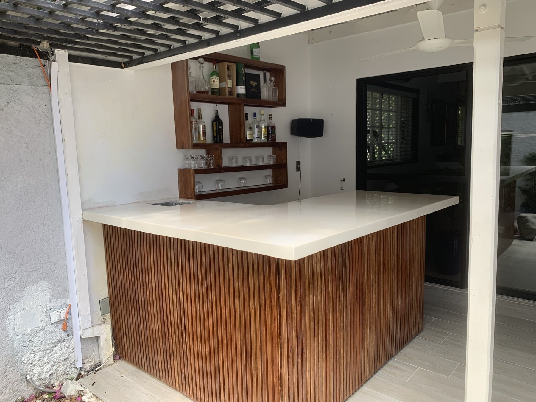 The complete L-shaped cream epoxy bar, with a fitted sink, timber-slatted base and stocked shelves beneath a pergola.
