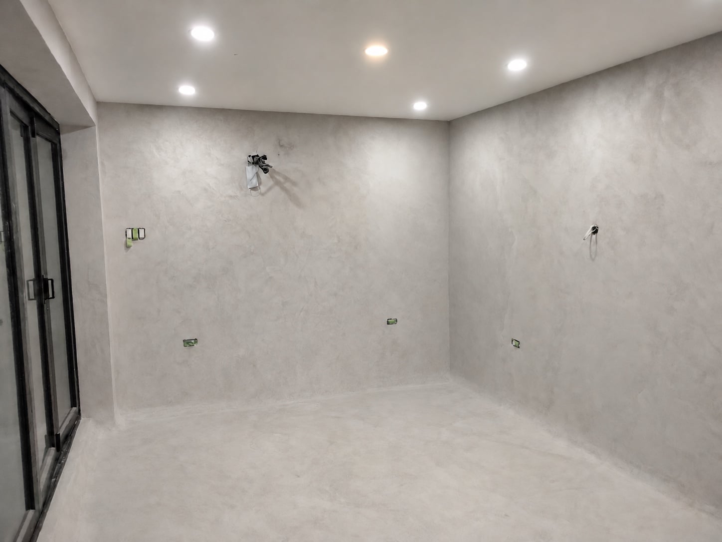 Retouched view of the pale-grey microcement sun room; the lower-wall tone and floor marks are blended while electrical fittings remain unfinished.
